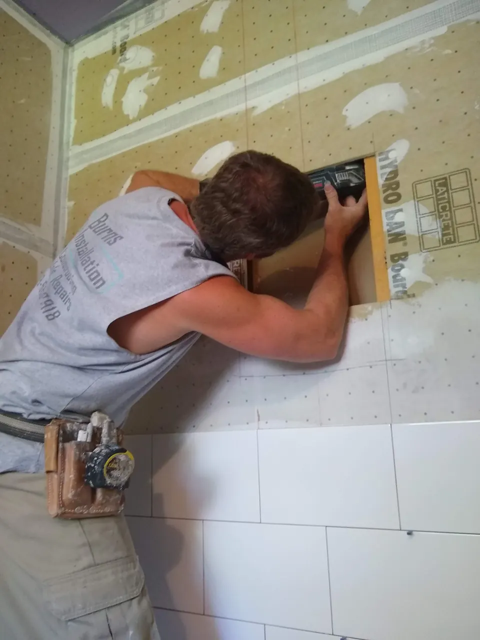 Tile installer working inside a niche opening in a shower wall of waterproof foam board, with large white tile set below